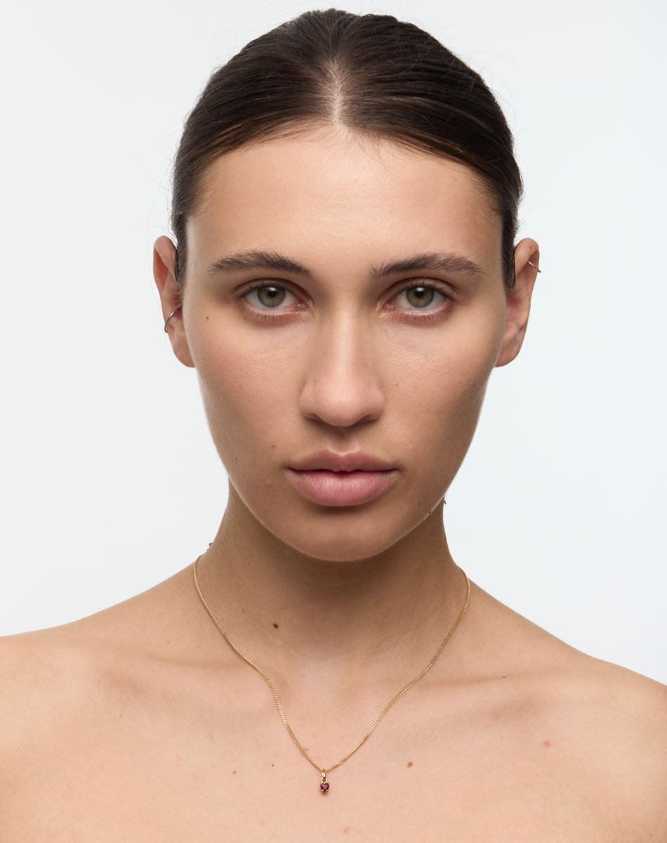 Woman with a plain background wearing a delicate necklace set with a red heart shaped gemstone.